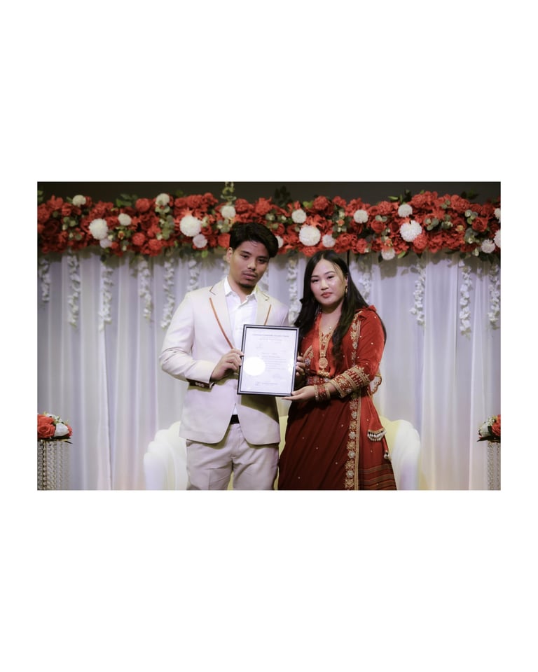 Bride and groom in traditional wedding attire holding a certificate in front of red and white floral backdrop