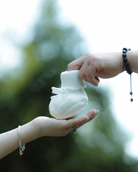 Two hands holding a pair of white baby socks outdoors with blurred green foliage in the background