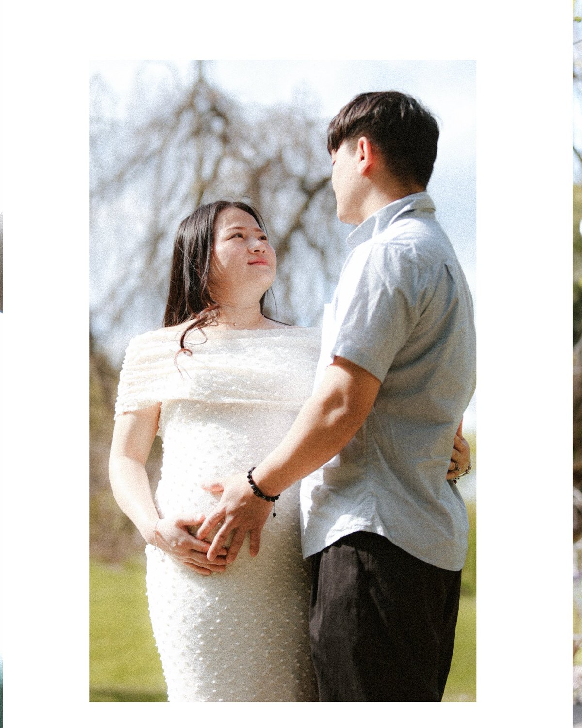 Pregnant couple standing outdoors, woman in white dress and man in light shirt holding her belly, bare trees in background