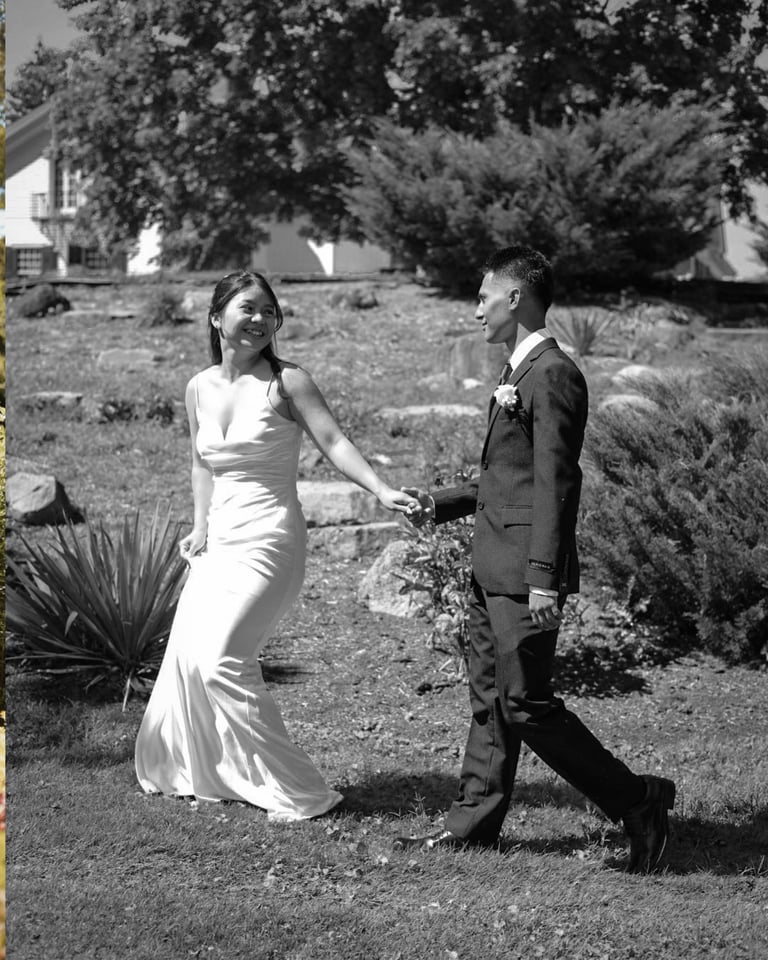 Black and white photo of a bride in a white gown and groom in a suit walking together in a garden setting