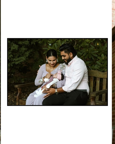 A couple sitting on a wooden bench outdoors, holding their newborn baby wrapped in white, with green foliage in the background