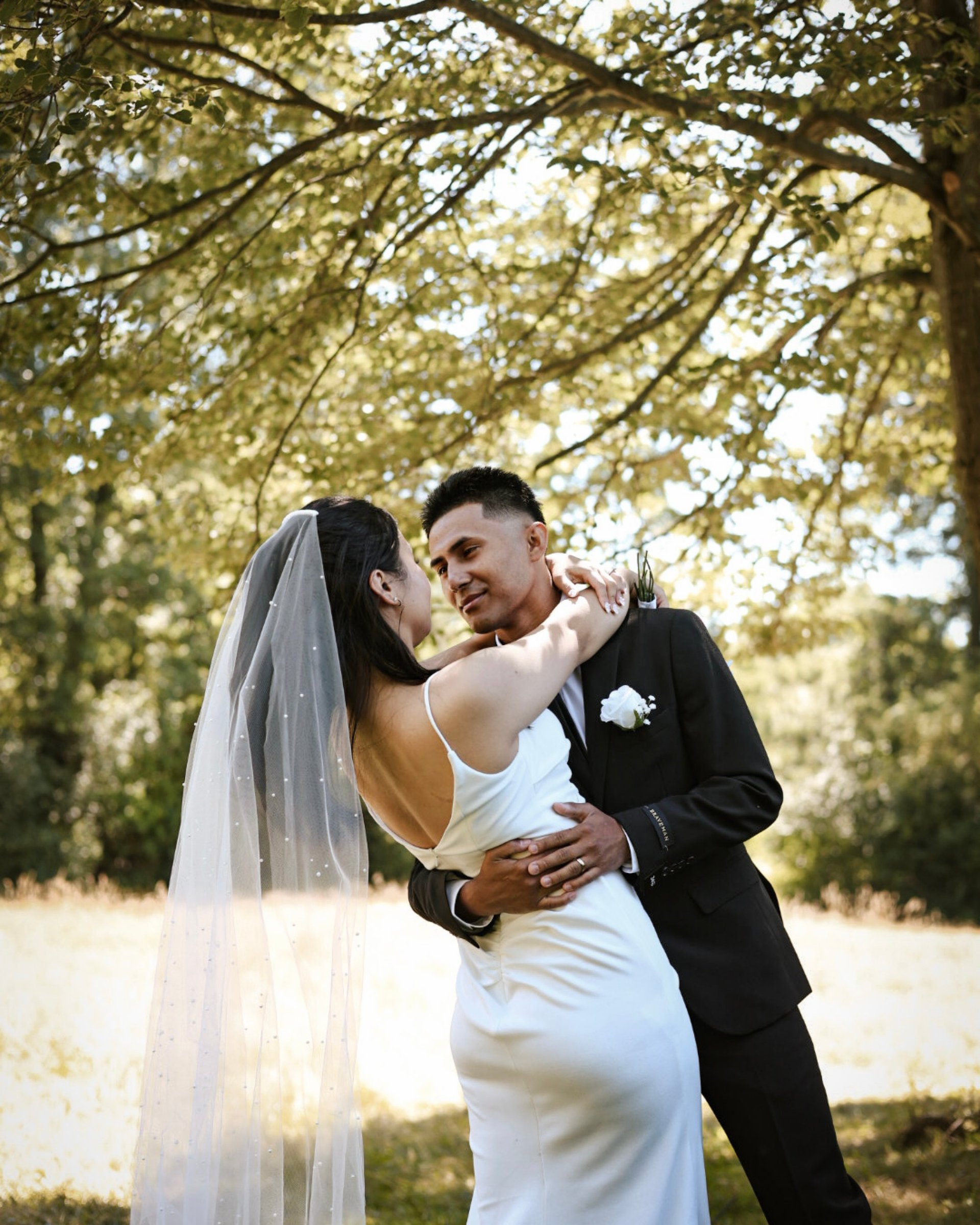 Newlyweds embracing under tree canopy on wedding day, bride in white dress and veil, groom in black suit