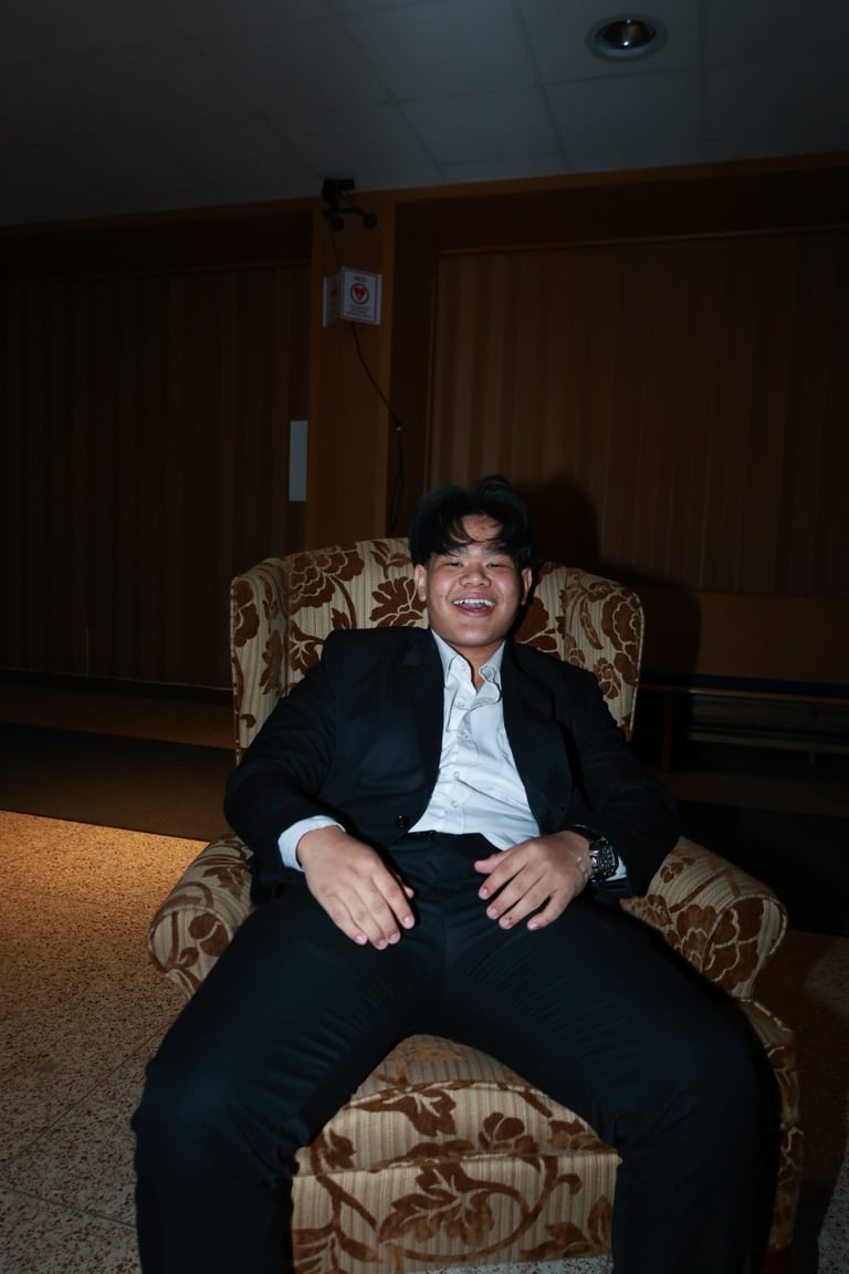 Man in dark suit and white shirt sitting on patterned armchair in home interior, smiling at camera